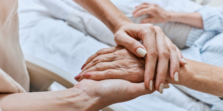 Two people holding hands in comfort and support, one is younger and the other is older and in a hospice bed - Tips for Handling Common Emotions During Hospice Care - Northern Illinois Hospice